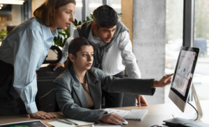 Three coworkers in an office review data on a large monitor as one woman points at the screen while two colleagues look on.