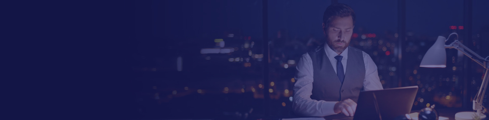 Bearded man in a gray vest and tie typing on a laptop at a night office with city lights outside a window.