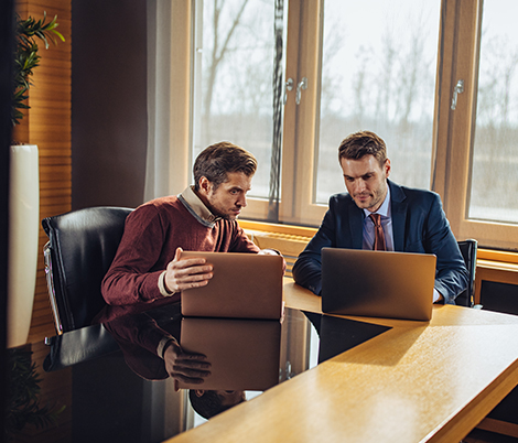 Two men in a bright office meet at a glass table, comparing laptops during a collaborative discussion by a windowed wall.
