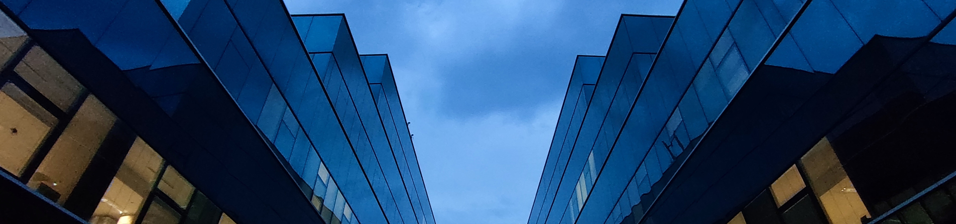Two blue glass office buildings converge toward the center against a cloudy sky, creating a V-shaped canyon.