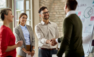 Group of four coworkers in a bright office shake hands while smiling near a whiteboard in the background.] ,