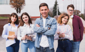 Five students stand together outdoors, smiling at the camera with books and notebooks in hand.