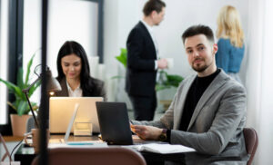 Man in a gray blazer sits at a desk with a laptop and papers, smiling at the camera; coworkers stand and chat in the background.