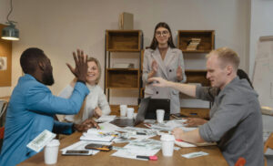 Diverse team around a conference table celebrate with high-fives, laptops, papers, and coffee cups visible.