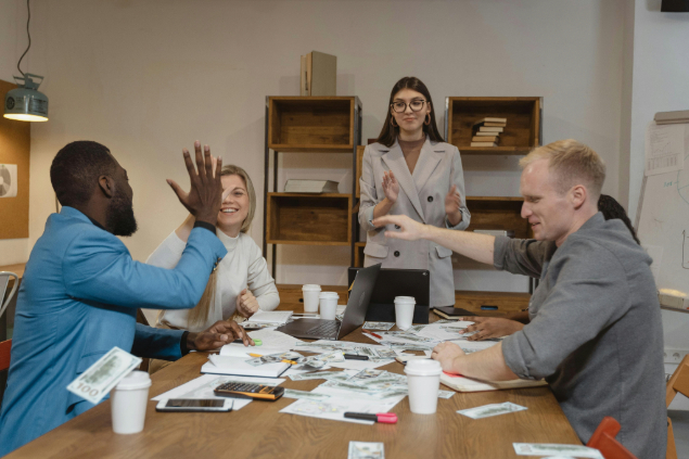 Diverse team around a conference table celebrate with high-fives, laptops, papers, and coffee cups visible.