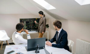 Business team in a bright attic-style office: two men sit at a white table with laptops and papers while a woman points to a monitor displaying charts and data.