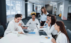 Six coworkers around a conference table with laptops and documents, collaborating in a bright open-office environment.
