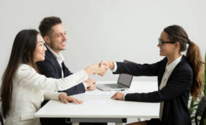 Two professionals across a white table shake hands as a laptop sits nearby, signaling a successful meeting or agreement.