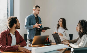 A team of four colleagues in a bright conference room; a man stands presenting with a clipboard while three others listen and use laptops or papers on the table.