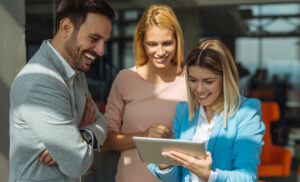 Three colleagues gather around a tablet, smiling as they review something together in a bright office.