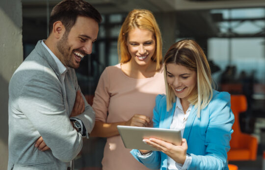 Three colleagues gather around a tablet, smiling as they review something together in a bright office.