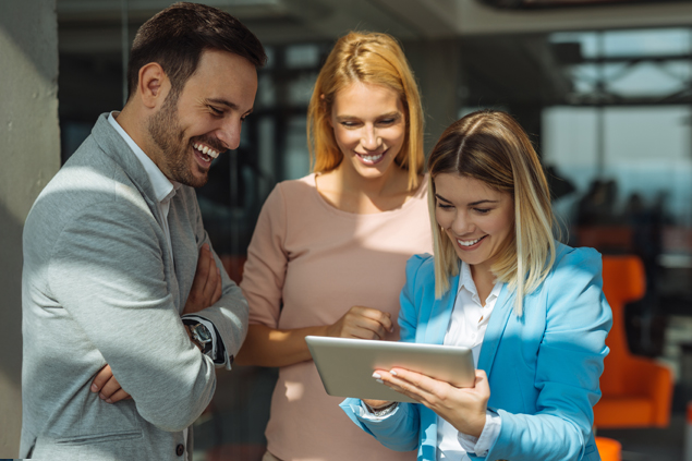 Three colleagues gather around a tablet, smiling as they review something together in a bright office.