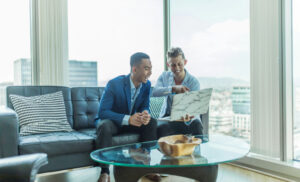 Two professionals in a bright office sit on a sofa, reviewing a large printout together near a window.