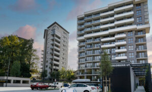 Modern residential complex with two tall apartment buildings, a parking lot, and greenery in a suburban setting.