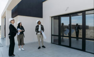 Four professionals wearing hard hats stand on a modern building terrace, two holding blueprints and examining the structure with glass doors nearby.