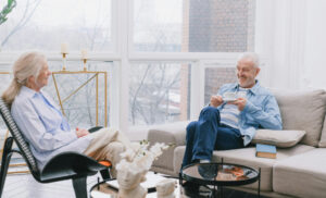 Senior couple relaxing in a bright living room; man holds a cup while chatting with the woman in a chair