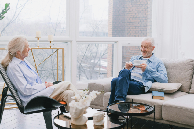 Senior couple relaxing in a bright living room; man holds a cup while chatting with the woman in a chair