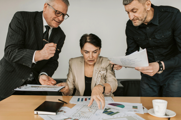 Three professionals review charts and documents on a conference table, one pointing at a graph as others look on.
