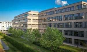 Modern brick office building with large windows, set in a sunny courtyard with trimmed hedges and trees in bloom.