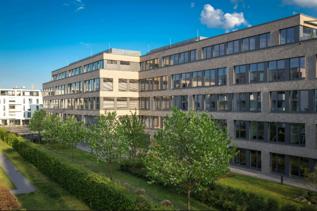 Modern brick office building with large windows, set in a sunny courtyard with trimmed hedges and trees in bloom.