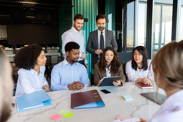 A diverse group of colleagues seated around a marble conference table, looking at a tablet together in a modern office.