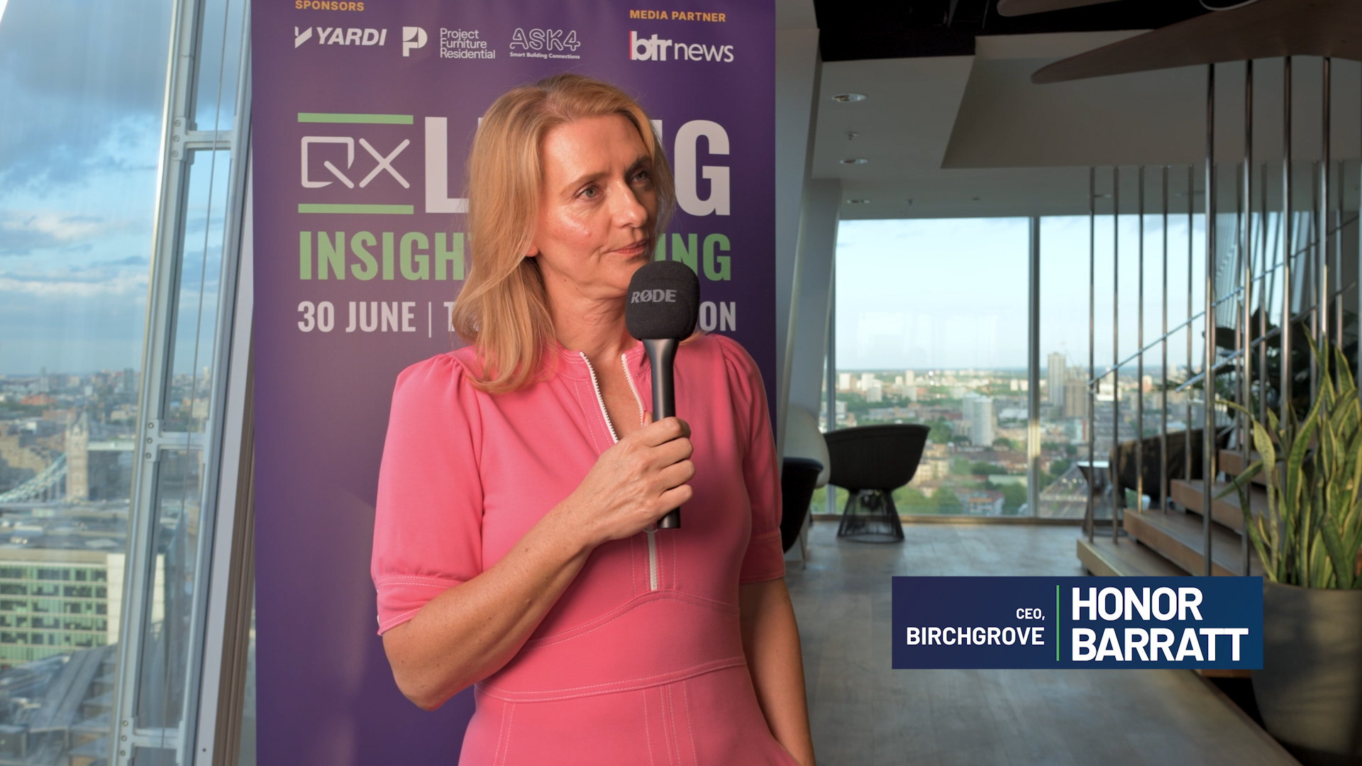 Woman in a pink dress holding a Rode microphone during an interview, with a city skyline visible through large windows behind her.