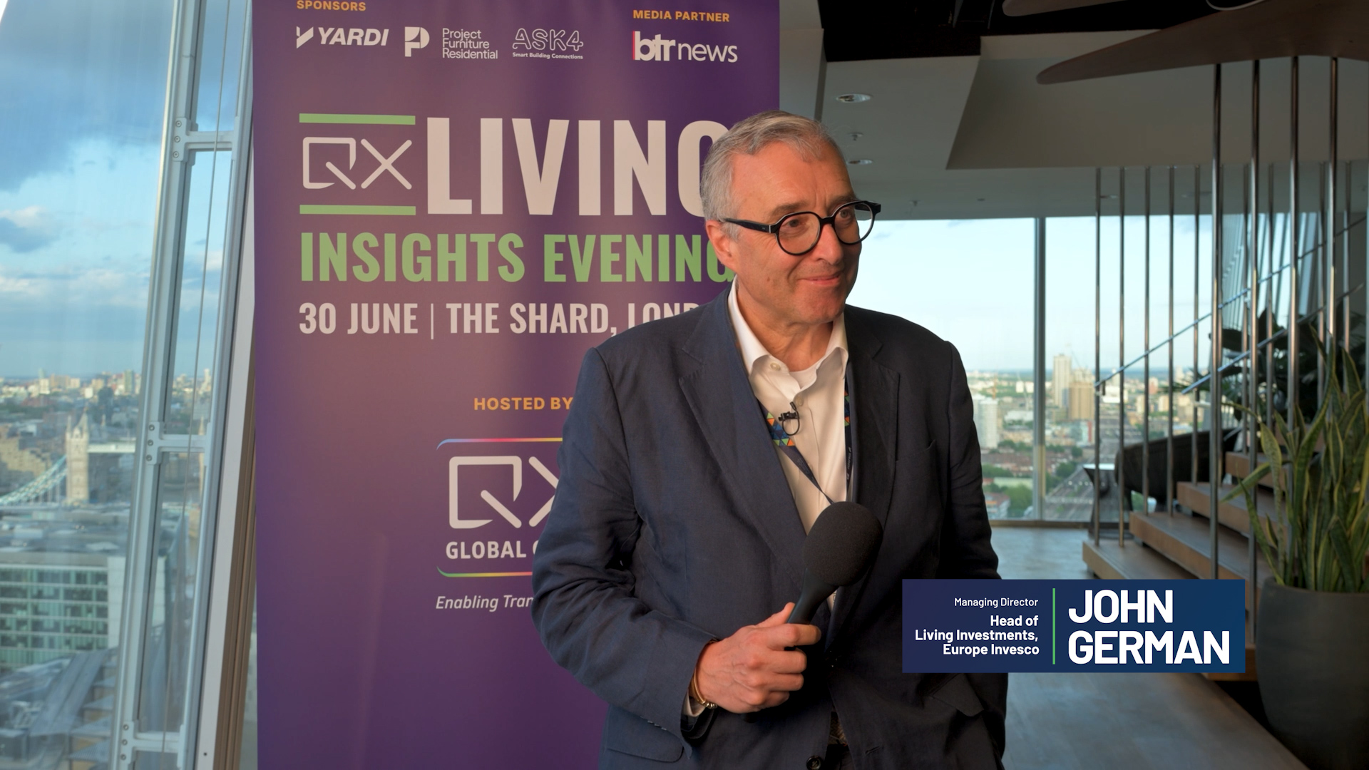 Man in a navy blazer and glasses being interviewed with a handheld microphone, standing beside a purple event banner reading 'LIVING INSIGHTS EVENING' against a city skyline backdrop.
