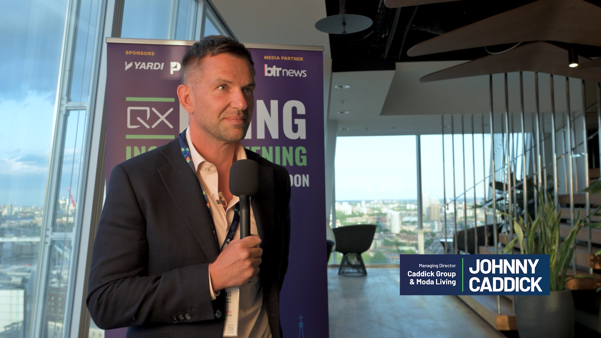 Man in a dark suit speaks into a handheld microphone at an event, with a purple banner behind him and city views in the background.