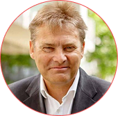 Portrait of a man with light hair in a dark suit jacket and white shirt, outdoors with greenery in the background.