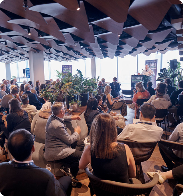 Audience seated in a modern conference room listening to a speaker at a podium with large windows and decorative ceiling panels nearby.