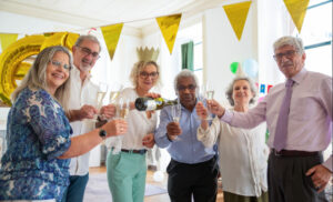 Six older adults toasting with champagne in a sunlit room, wearing party hats amid yellow bunting and balloons at a celebration.