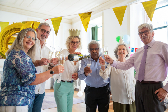 Six older adults toasting with champagne in a sunlit room, wearing party hats amid yellow bunting and balloons at a celebration.