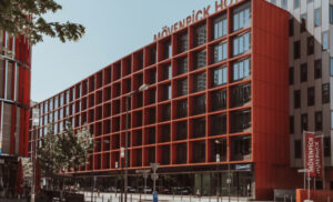 Red modern hotel building with a grid-like red facade labeled Mövenpick Hotel, seen from a street with trees, lampposts, and parked cars.