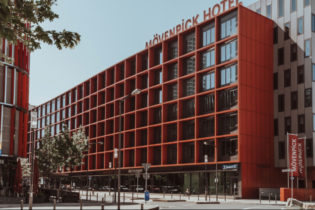 Red modern hotel building with a grid-like red facade labeled Mövenpick Hotel, seen from a street with trees, lampposts, and parked cars.