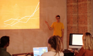 Presenter in a yellow shirt explains a rising line chart projected on a screen to a small audience in a workshop room with laptops nearby.