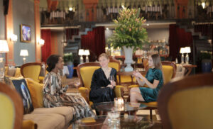 Three women in elegant dresses sit on yellow couches in a stylish lounge, chatting with wine glasses in hand.