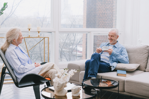 Two seniors chatting in a bright living room with large windows; woman sits in a chair, man on a sofa drinking tea.
