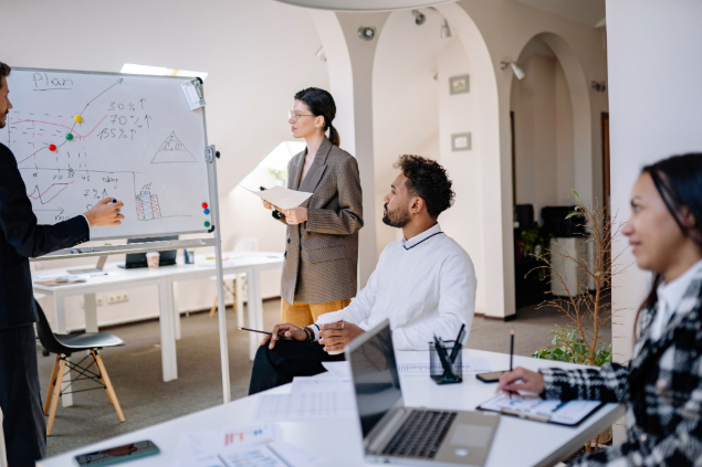 Team meeting in a bright office: a woman presents by a whiteboard with charts while a man sits and others work with laptops and papers.