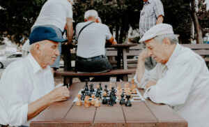 Two elderly men wearing caps sit across from each other at a wooden table playing chess in a park.