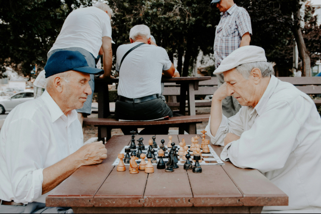 Two elderly men wearing caps sit across from each other at a wooden table playing chess in a park.