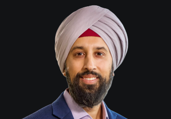 Portrait of a man with a light gray turban, beard, and a navy blazer, smiling against a dark background.