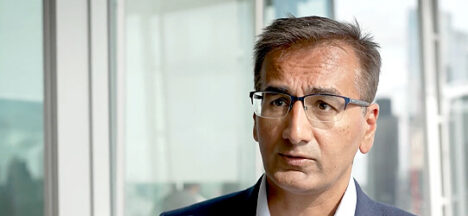Man in a suit and glasses looks to the side in a bright, modern office setting.