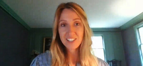 Smiling woman with blonde hair wearing a blue shirt, looking at the camera in a sunlit room with green walls and a window in the background.