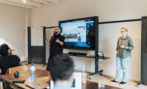 Two presenters lead a workshop in a bright room: a man speaks and gestures near a large monitor displaying content, while a woman stands to the side; attendees sit at tables listening.