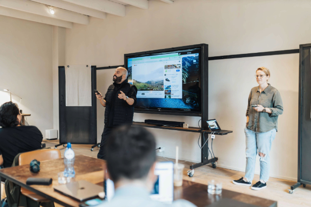 Two presenters lead a workshop in a bright room: a man speaks and gestures near a large monitor displaying content, while a woman stands to the side; attendees sit at tables listening.