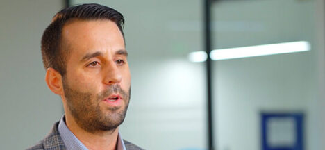 Close-up of a man with short dark hair and stubble speaking, wearing a gray blazer in an office setting.