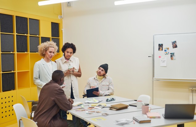 Four colleagues collaborate around a table in a bright office, with yellow storage units in the background.