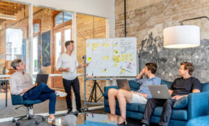 Man stands by a whiteboard presenting to three colleagues with laptops in a modern brick-walled office setting at a casual meeting table area on a sofa and chairs.