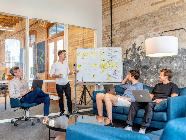 Man stands by a whiteboard presenting to three colleagues with laptops in a modern brick-walled office setting at a casual meeting table area on a sofa and chairs.