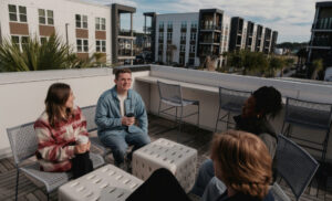 Group of four friends chatting on a rooftop terrace with drinks, modern apartment buildings in the background.